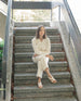 Woman sitting cross legged on stairs modeling Coclico Cardinal Sandal in Greige leather. 3