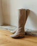Coclico Chip Boot in Limestone nubuck, a knee-high boot with elastic goring, seam detailing across arch, inside zip closure, and a low-height solid wood block heel - leaning against white wall on wooden flooring.  2