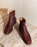 Coclico Egg Boot in Burgundy leather, a classic ankle boot with a flat round wood-heel, patch sole and an inside zip closure - on marble table.  3