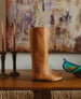 Coclico Haricot Boot in Luggage leather, a flat-heeled, knee-high boot with an inside zip closure and  seam detailing - in display with painting & decor in background.  2
