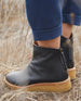 Close up side view of a woman's feet wearing the Hop Boot in Deep Sea while standing out wearing blue pants.  8