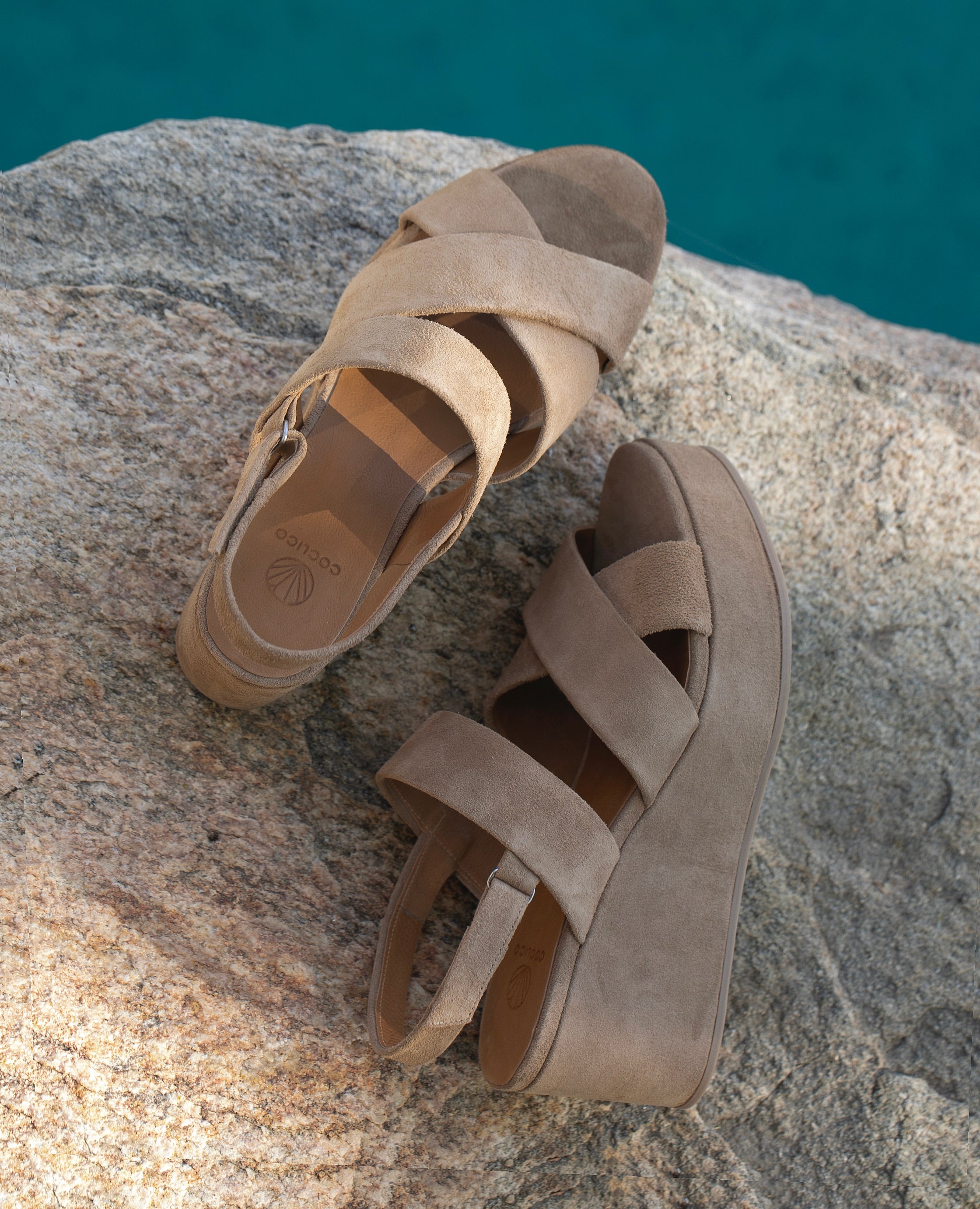 Close up of Coclico Lacine Wedge in Tobacco suede, angled view:a open crisscrossed sandal on a recycled cork wedge platform, back strap and velcro closure -  on a boulder by the pool.