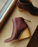 A sculpted 75mm solid wood wedge with a rounded toe and a back zip closure - Coclico Lovage Boot in Burgundy leather, displayed in side view on wooden window sill.  2