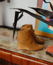 Side view of the Coclico Lovage Boot in Wheat suede: Sculpted 75mm solid wood wedge with a rounded toe and a back zip closure - placed on wood/marble table with decor in background.  4