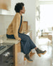 Model sitting on kitchen counter wearing a lace-up clog featuring a 3.5 inch solid wood heel, leather sole and inside zip closure - Coclico Vanji Clog in Anthracite suede.  4