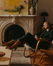 Model seated on low leather woven chair wearing the Wakame Boot in Mandorla leather, a sophisticated Coclico pointed-toe boot with a 50mm arched solid wood heel and inside zip closure - marble fireplace in background.  5