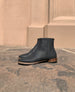 Outdoor, sunlit close up of the Coclico Egg Boot in Black Caviar textured leather: a classic ankle boot with a flat round wood-heel, patch sole and an inside zip closure. 1