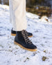 Women's legs in white pants and feet in the Heaven Shearling Boot in Black standing on snow. Angled view.  3