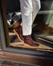 Legs of a woman standing in front of a glass door wearing white pants and the Coclico Hop Boot in Merlot.  7