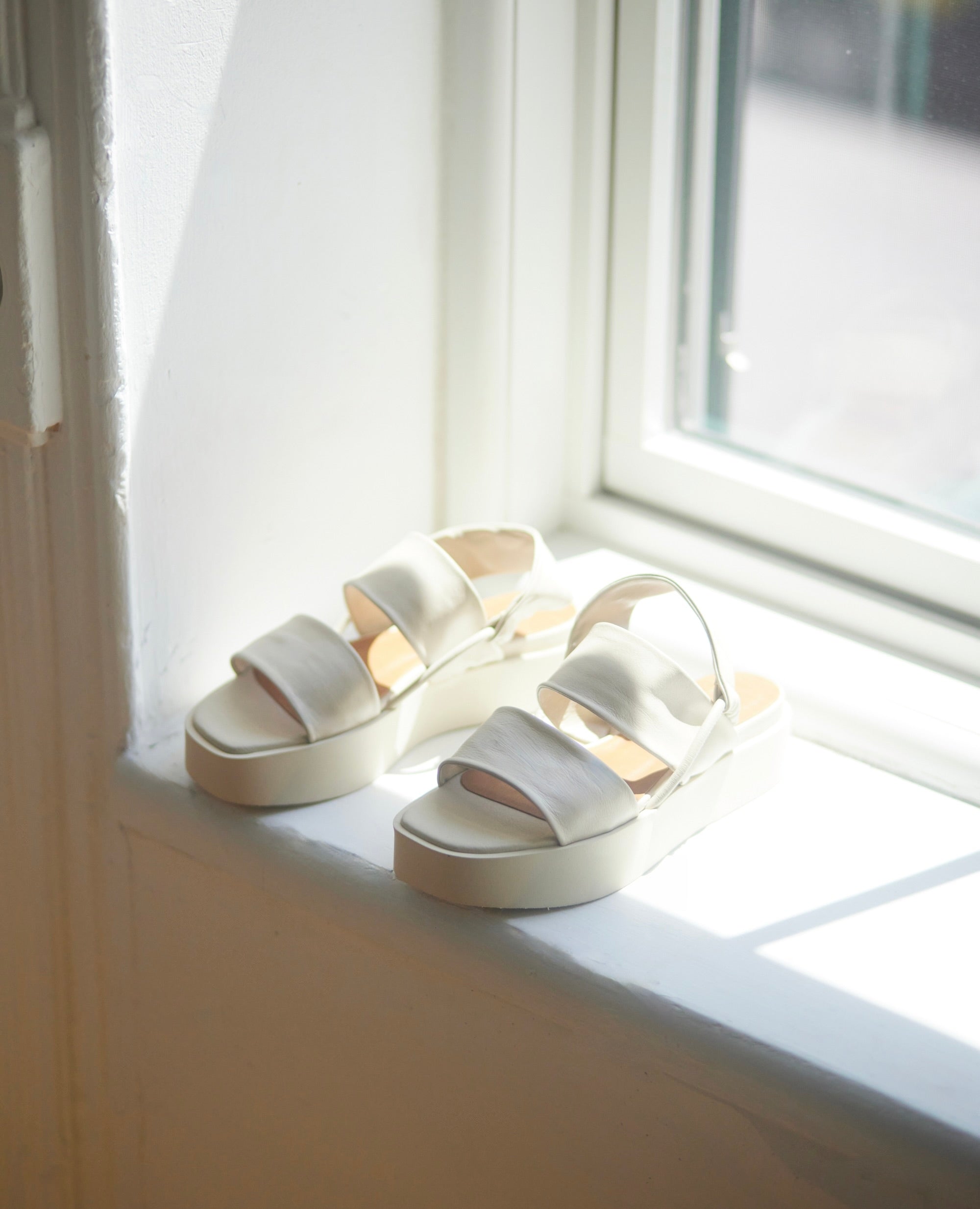 Close up angle view of an open wedge with 2 tubular straps across foot, mid-height EVA  flatform sole, elasticated slingback strap - placed on window sill in sunlight.