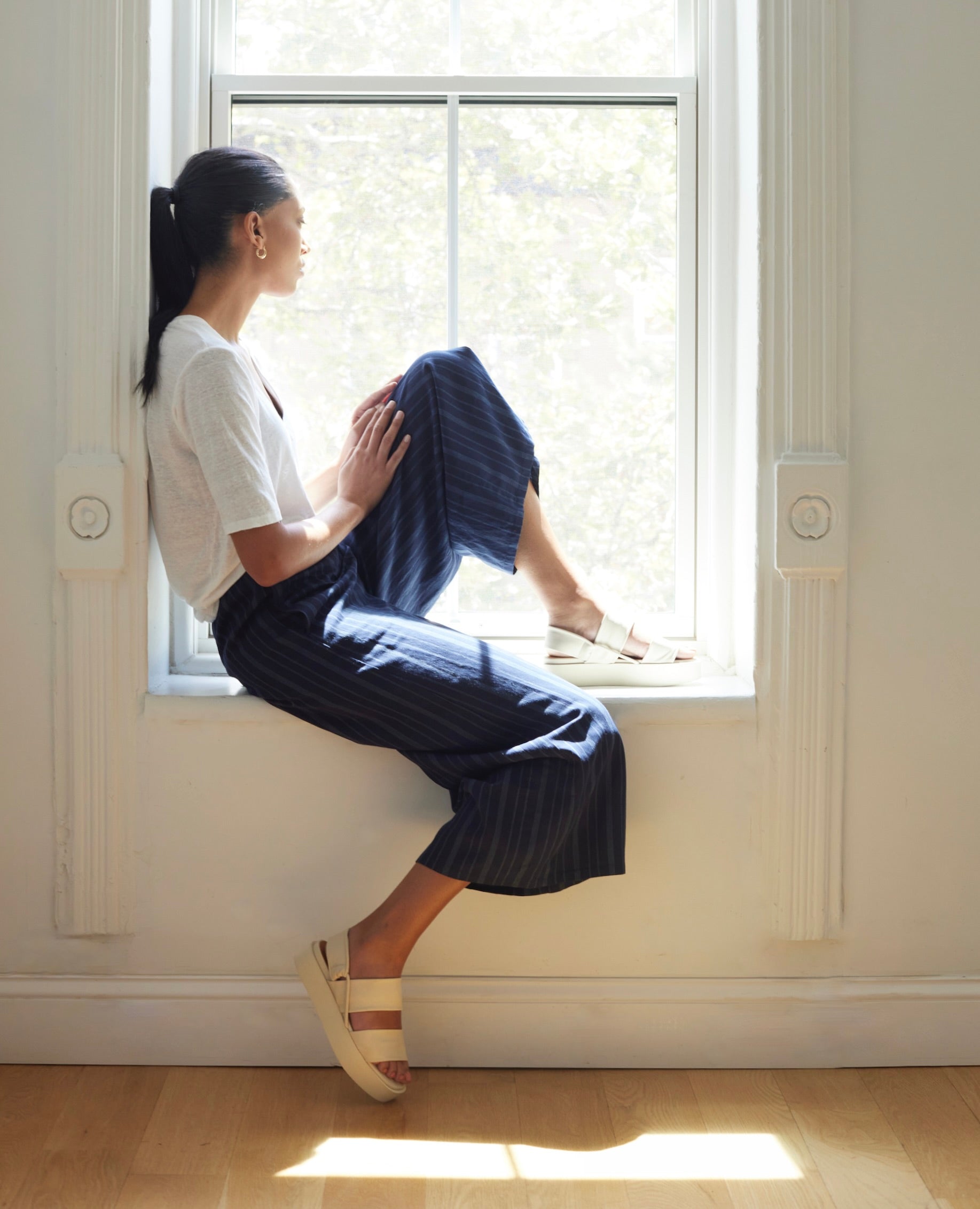 Woman sitting on window sill in sunlight modeling Coclico Imna Wedge in Greige leather: open wedge with 2 tubular straps across foot, mid-height EVA  flatform sole, elasticated slingback strap.