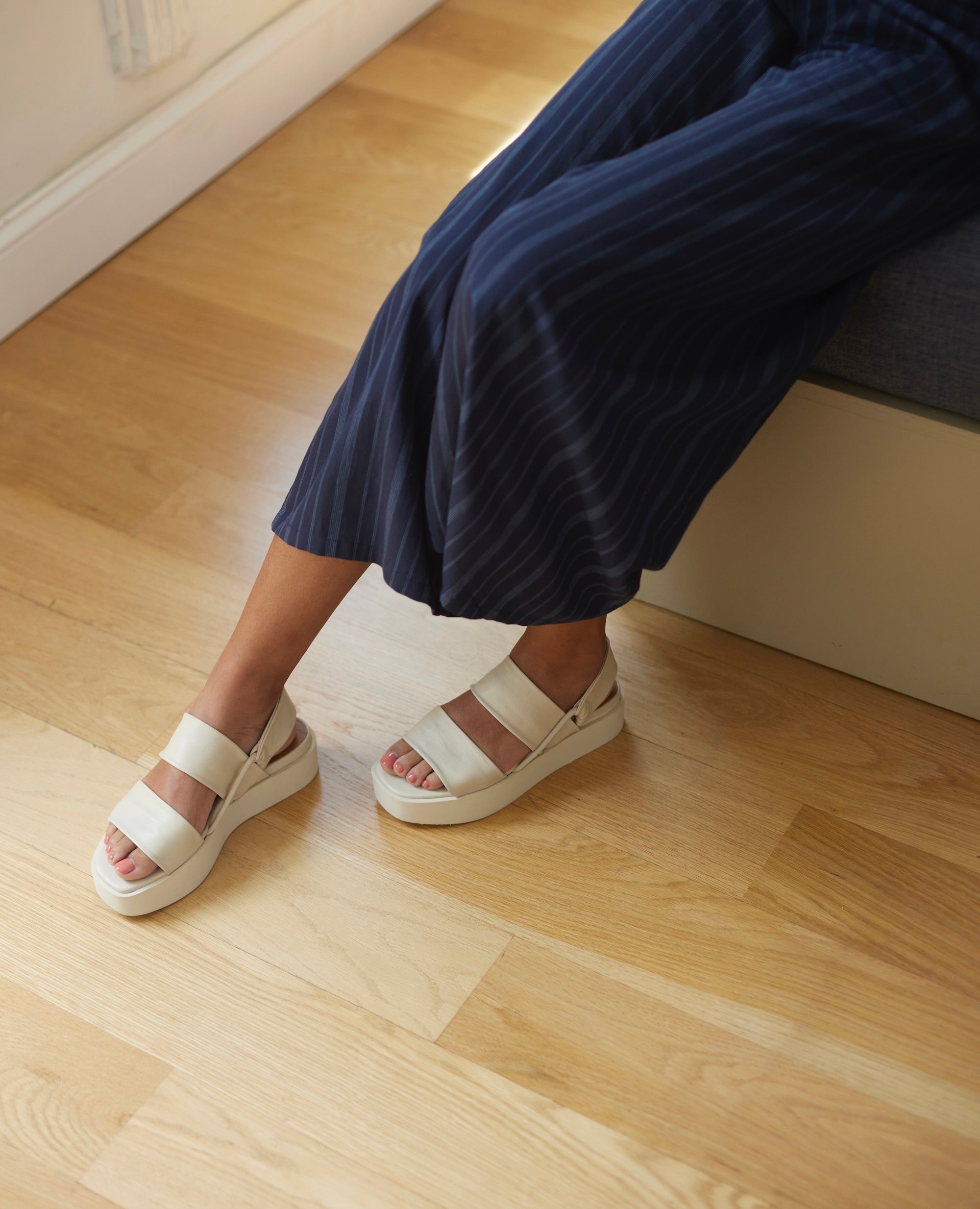 Model sitting on edge of bed wearing Coclico Imna Wedge in Greige leather: open wedge with 2 tubular straps across foot, elasticated slingback strap, mid-height EVA flatform sole.