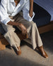 Lower half of a woman sitting on a bed resting her elbows on her legs wearing a white button up shirt with beige pants and the Ione Boot in Caramelllo. 3