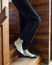 Legs and feet of a woman wearing blue corduroy pants with the Krispin High Top Sneaker in Griege Nubuck standing on a wooden staircase.  2