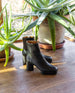 The Travis Boot in Black placed on a wooden floor with plants in the background.  8