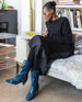 Woman seated on a couch reading a book and wearing the Wakame Boot in Painter's Blue.  5