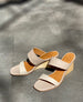 Coclico Doe Wedge in Greige & Marmo leather placed on tile flooring with natural sunlight: Doe Wedge, a two-neutral-toned slide, asymmetric straps, solid wood mid-height wedge. 7