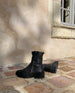 Side back view of Durum Shearling Boot on brick floor. This black boot is displayed at front angled view to show its full shearling interierior, outside zip encolusre, and updated upper lug sole. The right black boot lying forward to display the upper lug sole 6