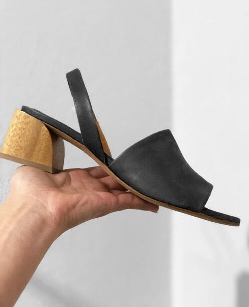 Woman presenting in hand the Coclico Okolo Sandal in Black leather, angled view: slingback sandal with wide stretch leather band across the foot and softly curved, low, solid wood heel. 