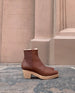 Side view close up of the Coclico Keep Shearling Clog Caramello leather, a shearling-lined clog with tapered, snipped toe, inside zip closure, mid-height wood block platform - with terracotta wall in background. 1