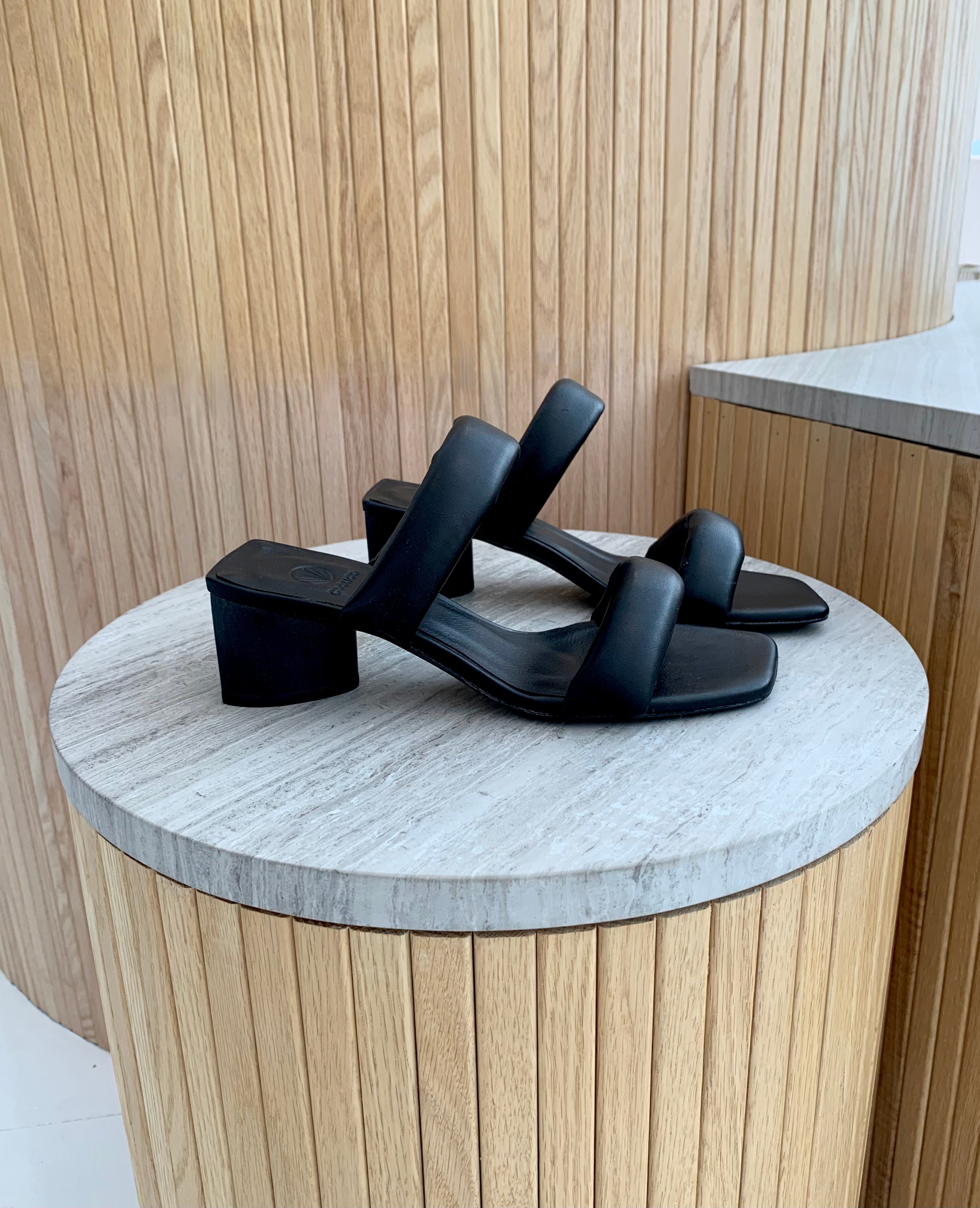 side view of a mid-heel all black slide sandal with two padded straps across the foot. Sows the flat back of the wood heel.