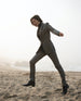 Woman wearing a black turtleneck with a grey suit and the Midori Bootie in Black skipping on a beach.  6