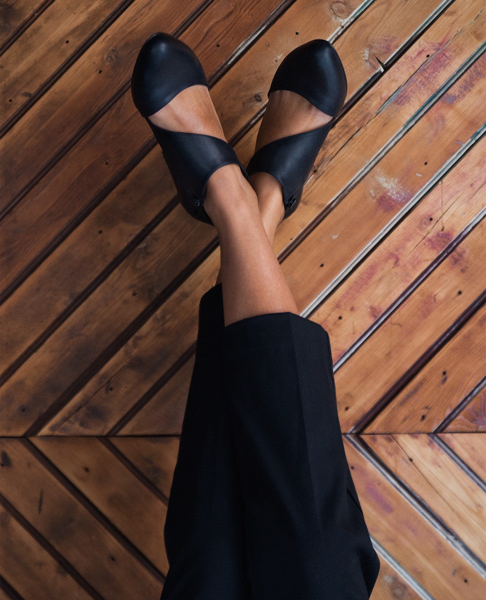 Woman seated crossed legged modeling the Coclico Sarah Heel in Black leather, a closed-toe mid-heel pump, with an asymmetrical opening and zip closure. 