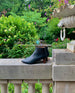 The Coclico Whoop Boot in Deep Sea leather, placed on a stone wall in a flourishing garden. 6