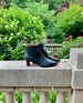 A timeless day-to-night ankle bootie with a pointed toe, 1.5 inch scultped solid wood heel and inside zip closure - side view with greenery in background.  7