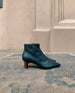 Side view of a timeless day-to-night ankle bootie with a pointed toe, 1.5 inch scultped solid wood heel and inside zip closure  - outdoor shot on stone ground with terracotta colored building in background. 5