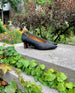 The Coclico Wumo Heel in Black/Coabi leather, a seasonless slip-on heel with a choked throat line, pointed toe, scultped oval shaped solid wood heel and subtle stichwork - placed on stone block with greenery in background. 6