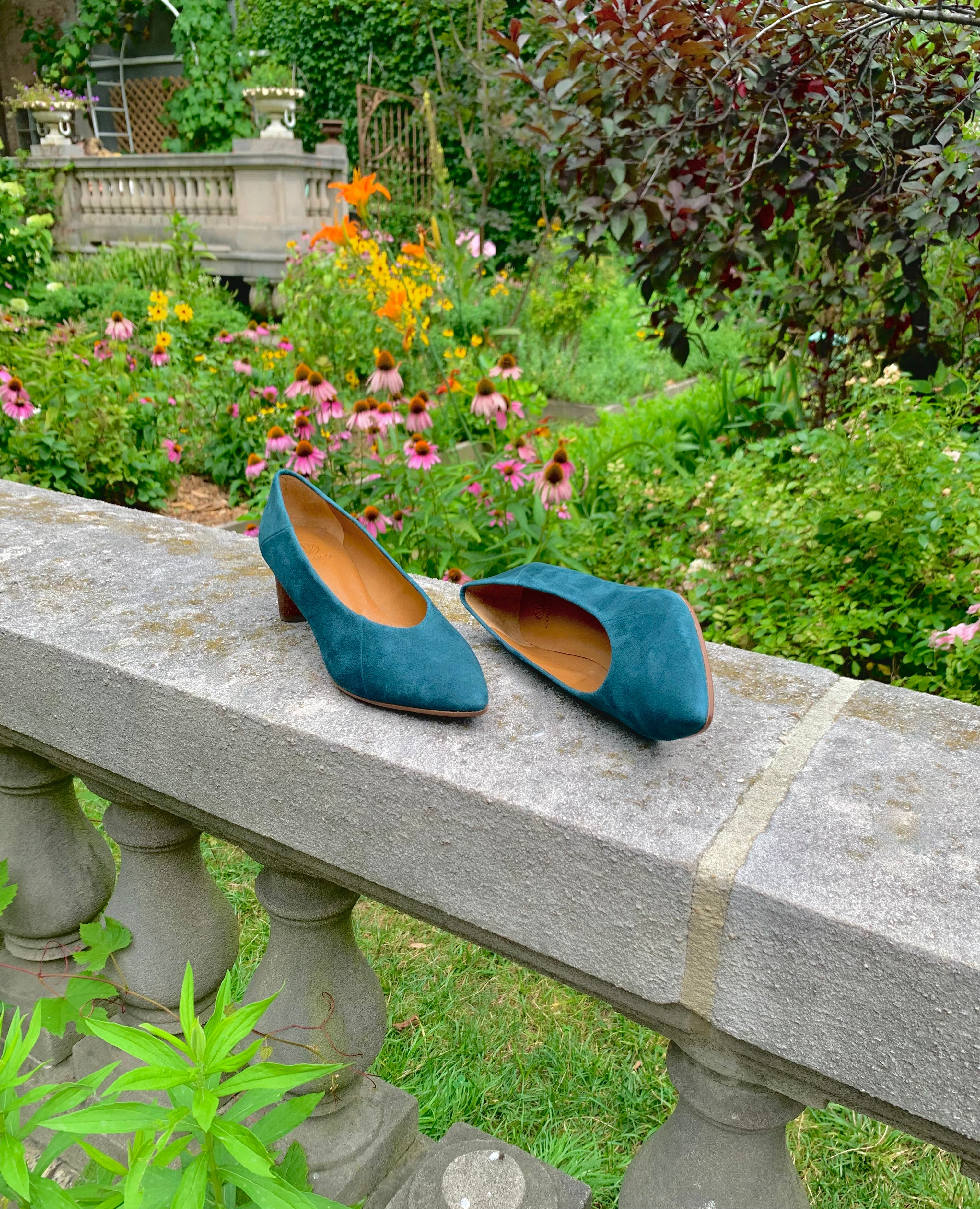Coclico Wumo Heel in Bottle suede with a scultped, oval shaped solid wood heel in a hand-painted chestnut finish - placed on stone wall with greenery in background.
