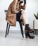 Woman sitting on a chair wearing a beige coat with her legs crossed, and feet wearing the Colico Tecla Clog in Black.  4